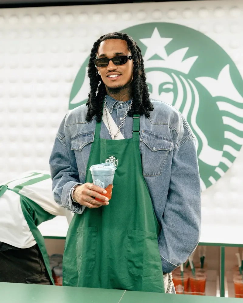 A guest barista in a green Starbucks apron holding an iced drink